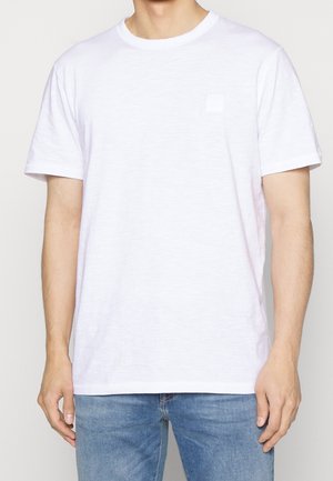 Mand iført almindelig hvid kortærmet t-shirt med rund hals og blå jeans, stående med armene afslappet ned langs siden, mod en lys baggrund.