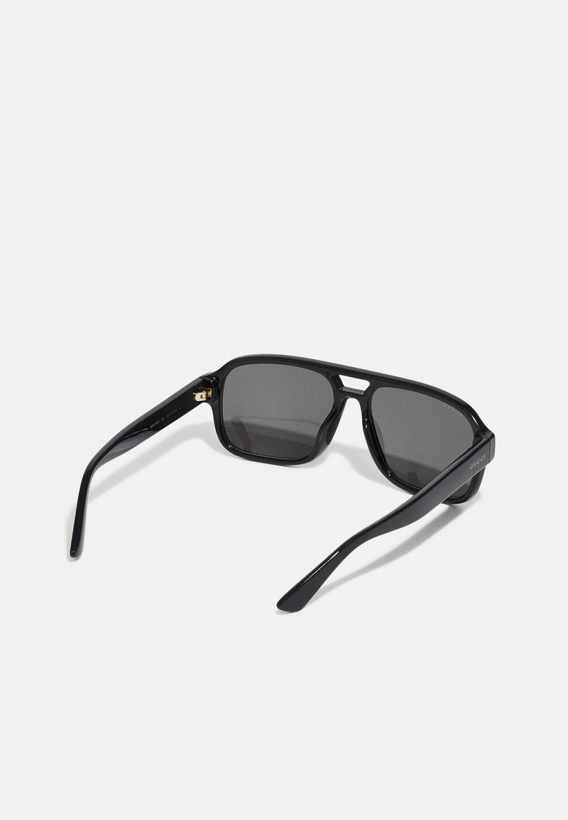 Lunettes de soleil rectangulaires noires avec des verres sombres, finition brillante, branches étroites et détails dorés subtils sur le côté. Design minimaliste.