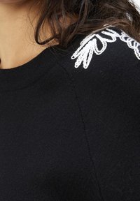 Suéter de punto negro con cuello redondo, tela texturizada y bordado floral blanco en el hombro como detalle.