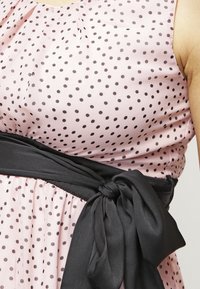 Bezramínkov dress v světle růžové barvě s černými puntíky, s černým páskem z látky uvázaným v pase, vytvářející strukturovanou siluetu.