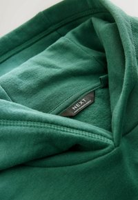 Grön hoodie med ett mjukt, texturerat tyg. Har en dragsko i huvan och en etikett som säger "NEXT, tillverkad i Bangladesh."