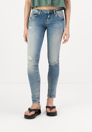Jean skinny bleu avec un délavage et des détails usés. Porté avec un haut vert court et des sandales à plateforme noires.