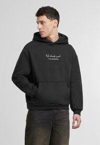 Crna hoodie dukserica napravljena od pamučne mješavine. Sadrži prednju džep i bijeli natpis: "أحب نفسك أولاً - Voli sebe prvo." Jednostavan dizajn.