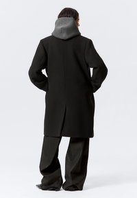 Άτομο που φοράει μακρύ μαύρο παλτό πάνω από γκρι φούτερ με κουκούλα και φαρδιά μαύρη παντελόνια, στέκεται με τα χέρια στις τσέπες, στραμμένο με την πλάτη στην κάμερα.