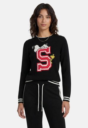 Frau trägt einen schwarzen Pullover mit einem großen roten Buchstaben S und den Comicfiguren Snoopy und Woodstock, kombiniert mit schwarzen Jogginghosen mit weißen Streifen.