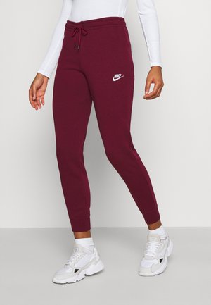 Kobieta w bordowych spodniach dresowych Nike z białym logo, białej bluzce z długim rękawem, białych skarpetach i białych butach sportowych stojąca na szarej podłodze.