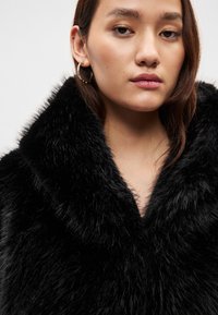 Juodas dirbtinio kailio paltas su plačiu apykakliu, pasižymintis minkštu tekstūra ir gilia V formos iškirpte. Modelis demonstruoja minimalias aksesuarus.