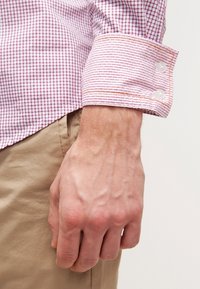 Mano masculina descansando junto a pantalones kaki, con una camisa de manga larga a cuadros rojos y blancos y puño abotonado.