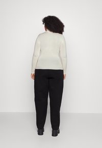 Top a maniche lunghe beige chiaro abbinato a pantaloni neri larghi. Texture liscia, forma aderente e design minimalista. Calzature nere completano l'outfit.