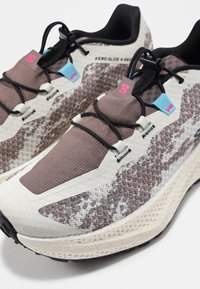 Gesloten veters hardloopschoenen met beige, bruin en zwart patroon op het bovenwerk, dikke witte zolen en roze en blauwe lusjes met het label "S".