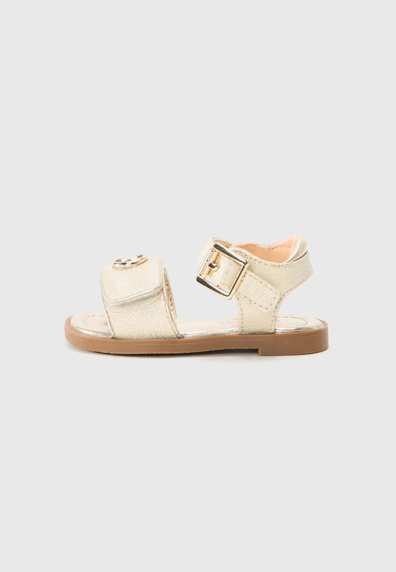 Kultaiset metalliset sandaalit, joissa on litteä pohja, solkikiristys ja koristeellinen pyöreä aksentti yläremmissä. Sileä pinta.