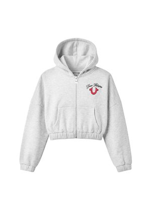 Femme portant un sweat à capuche court gris zippé et un pantalon de jogging large assorti avec un logo rouge, des baskets blanches, des lunettes de soleil et des boucles d'oreilles créoles.