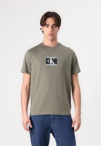 Olivovo zelené bavlnené tričko s okr�úhlym výstrihom, krátkymi rukávmi a obdlžníkovým čiernym logo grafickým dizajnom obsahujúcim "Calvin Klein" a "cK".