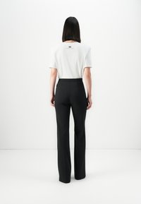 Woman with straight dark hair wearing a white short-sleeve top and high-waisted black trousers standing against a plain white background.