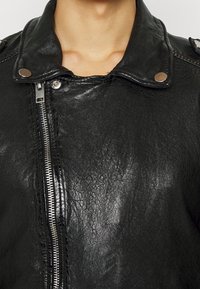 Czarna skórzana kurtka z przednim zapięciem na zamek, kołnierzem zapinanym na napy oraz teksturowaną powierzchnią na górnej części ciała osoby.