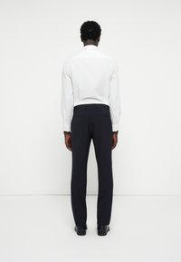 Weißes Hemd mit langen Ärmeln und Knopfleiste, kombiniert mit einer dunklen Hose. Das Hemd hat einen klassischen Kragen; die Hose ist schmal geschnitten und hat eine gerade Beinform.