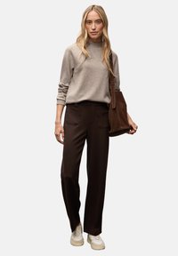 Un pull gris clair à col rond associé à un pantalon large marron foncé. Le mannequin porte un sac à main en daim marron foncé. Des baskets blanches complètent la tenue.