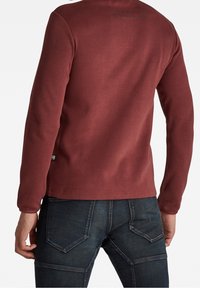 Chemise à manches longues bordeaux vue de dos, en coton avec une texture lisse. Présente un col rond et un logo discret près du col.