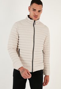 Lys beige pufferjakke med horisontale syninger, høj krave og sort lynlås. Har frontlommer og en glat, mat overflade.