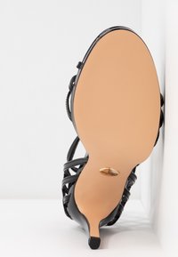 Sandale noire à talons hauts avec semelle beige, fines lanières et un petit logo doré près du talon, photographiée par en dessous sur fond blanc.