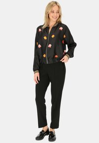 Czarna satynowa kurtka bomberka z kwiatowym haftem w odcieniach różu i pomarańczu, z ściągaczami przy mankietach i zamkiem z przodu, w połączeniu z dopasowanymi czarnymi spodniami i błyszczącymi butami.