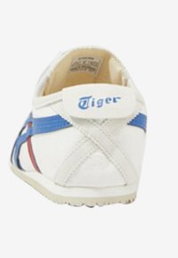 Sneaker bianca con accenti blu e rossi. Presenta un design con cinturino incrociato e un'etichetta logo prominente sul tallone. Suola in gomma.