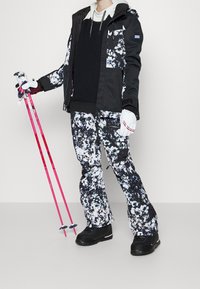 Person i svart och vit blommig skidjacka och byxor, svarta stövlar, vita handskar, som håller två rosa skistavar.