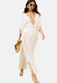 Weißer, leichter Kimono-Style Überwurf mit Quastenbesatz und Spitzenverzierungen. Kombiniert mit einem beigen Bikini und einer geflochtenen Handtasche.