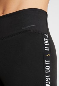 Detailní záběr na černý sportovní pás u legín s logem Nike a textem "JUST DO IT" na bočním švu.