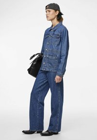 Giacca di denim con due tasche frontali, bottoni e colletto, abbinata a jeans a gamba larga. Borsa nera con manico decorato. Cappello indossato al contrario.