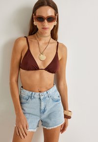 Top de bikini triangular marrón, shorts de mezclilla desgastados en azul claro con dobladillo desflecado, collar de conchas dorado, gafas de sol y pendientes en forma de concha.