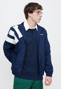 Tengerészkék széldzseki fehér átlós csíkokkal az ujjain, elöl cipzáros, rugalmas mandzsettákkal, sötét póló és zöld rövidnadrág fölött viselve.