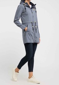 Femme portant une veste à capuche zippée grise avec cordons, un jean skinny foncé et des baskets blanches, se tenant avec une main dans la poche.