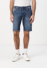 Shorts en denim bleu foncé avec une coupe droite, dotés de cinq poches, d'un léger effet délavé et d'ourlets retroussés. Associés à des baskets blanches.