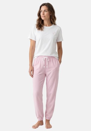 Vrouw staat blootsvoets met een wit T-shirt met korte mouwen met een klein glimlachend gezichtje en lichtroze joggingbroek met trekkoord tegen een effen achtergrond.