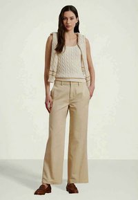 Mulher a usar um top sem mangas de malha em bege com um suéter correspondente pousado nos ombros, calças largas em bege e loafers castanhos.