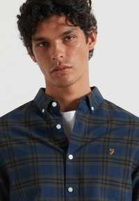Jonge man met krullend donker haar, gekleed in een blauw-groene geruite overhemd met een geborduurd bruin "F" op de borst.