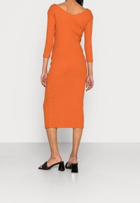 Robe midi orange côtelée avec des manches trois-quarts, décolleté en V et fentes latérales ; associée à des sandales noires à talons carrés.