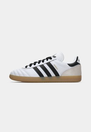 Weiße Adidas Samba Sneaker mit schwarzen Streifen, schwarzen Schnürsenkeln, beigem Wildleder-Fersenbereich und Gummiaußensohle, gezeigt im Profil von der linken Seite.
