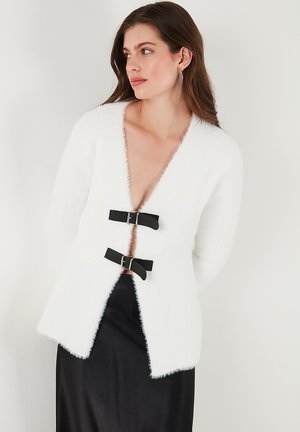 White fluffy cardigan with a deep V-neck, featuring two black rectangular buckle clasps at the front and long sleeves. Smoothed texture.