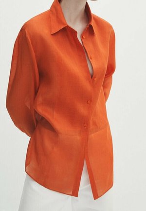 Vrouw draagt een doorschijnend oranje, lang mouw knoopoverhemd over een witte broek, met handen achter de rug op een effen witte achtergrond.