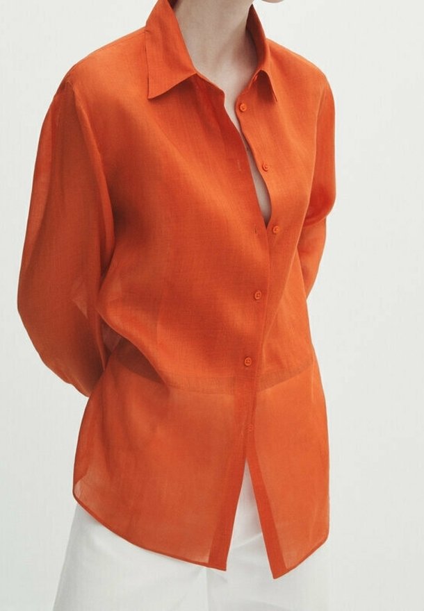 Femme portant une chemise orange transparente à manches longues boutonnée par-dessus un pantalon blanc, les mains derrière le dos sur un fond blanc uni.