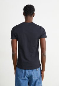 Tricou negru din bumbac cu mâneci scurte, de tip crew neck, cu un detaliu subtil de aplicație pe spate, purtat cu pantaloni din denim albastri.