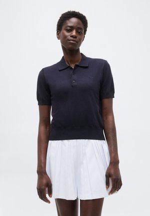 Žena oblečená v tmavomodrom polokošeli s krátkym rukávom a bielej plisovanej sukni, stojaca pred jednoduchým svetlým pozadím.