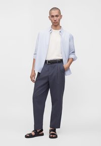 Homme portant une chemise rayée bleu clair à boutons sur un t-shirt blanc avec un pantalon gris foncé et des sandales noires, debout avec une main dans la poche.