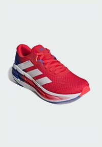 Roter Sportschuh mit Mesh-Obermaterial, weißen Streifen und lila Akzenten. Verfügt über eine gepolsterte Sohle und eine strukturierte Zehenkappe. Leichtes Design.