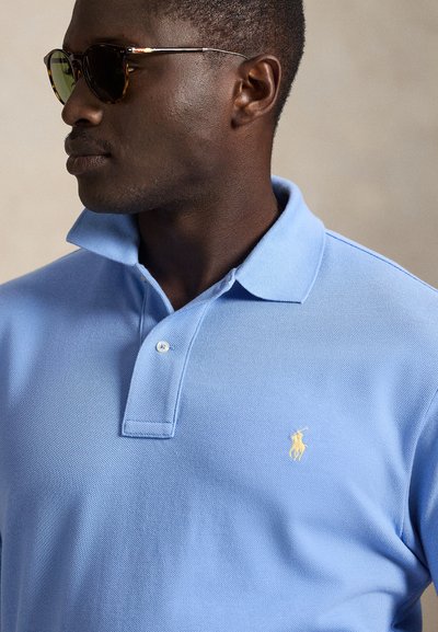 Ανοιχτό μπλε πόλο πουκάμισο με υφασμάτινη υφή, διαθέτει πατιλέτα με δύο κουμπιά και μικρό κίτρινο λογότυπο στο αριστερό στήθος. Το μοντέλο φορά σκούρα γυαλιά ηλίου.