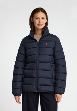 Donna che indossa una giacca imbottita trapuntata blu navy con un piccolo logo rosso sul petto, in piedi davanti a uno sfondo chiaro e uniforme.