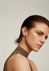 Top marrón de cuello alto con un acabado suave. Cabello corto y oscuro, peinado hacia atrás. Primer plano del rostro con un maquillaje sutil y aretes de perno.