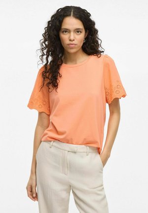 Top de algodón suave de color coral con mangas cortas y detalles de encaje cortado con láser en las mangas, combinado con pantalones de corte crema.